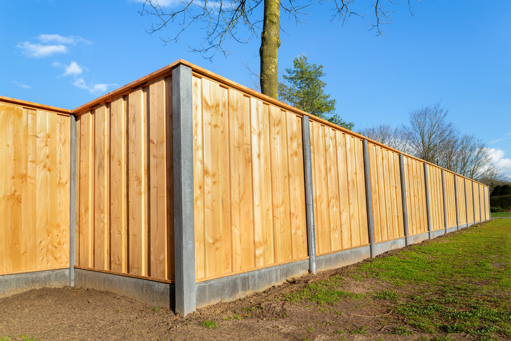 Brand new outdoor wooden fence, receding into distance, framed by blue sky, bright green grass, dirt and tree cover.