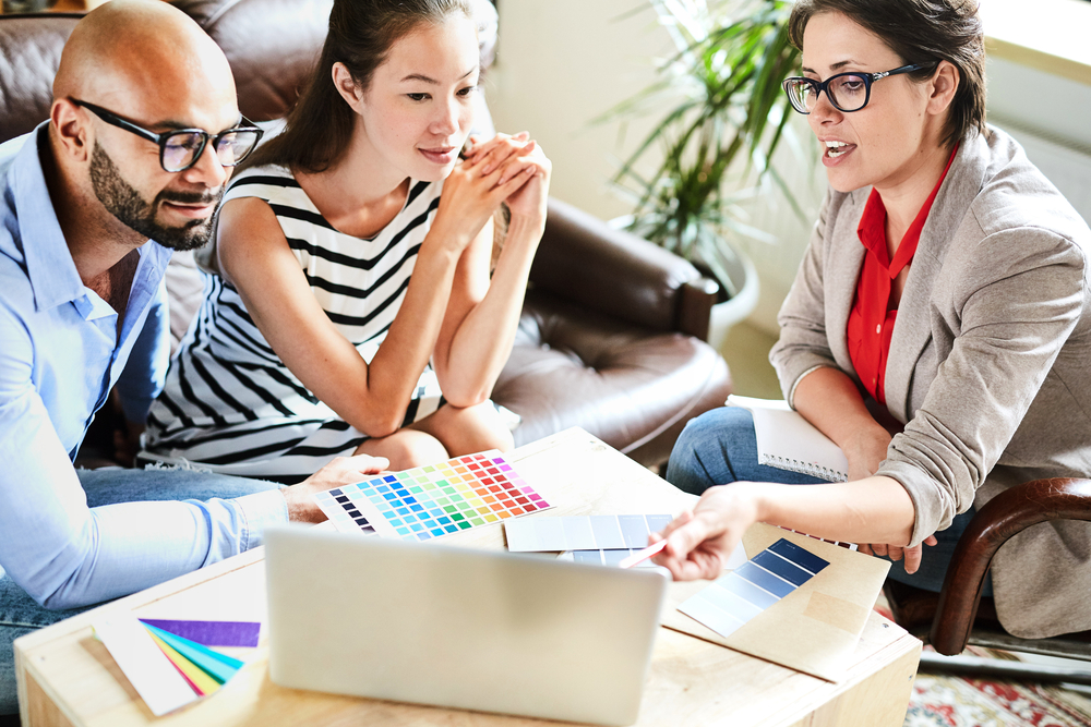 Designer pointing to computer at table with pens and palettes, showing young couple something on screen.